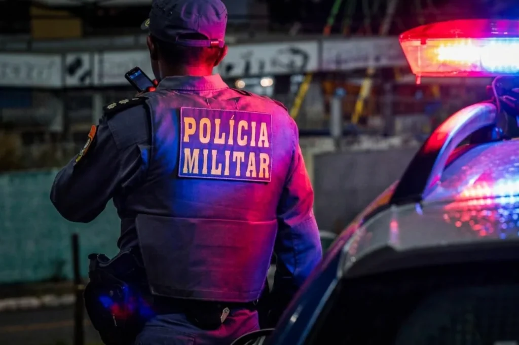 Policial militar ao lado de viatura com as luzes da sirene ligadas