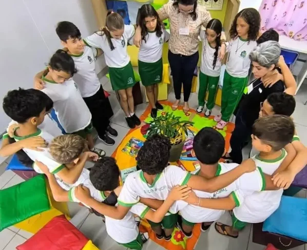 Foto vertical que mostra várias crianças e duas professoras abraçados em círculo, durante a participação em um círculo de construção de paz.