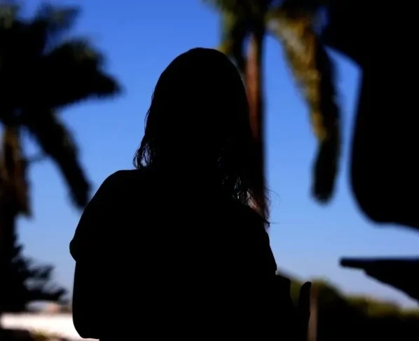 Foto horizontal que mostra a silhueta (sombra) de uma mulher vítima de violência doméstica. Ao fundo, aparece um céu azul e as sombras de coqueiros.