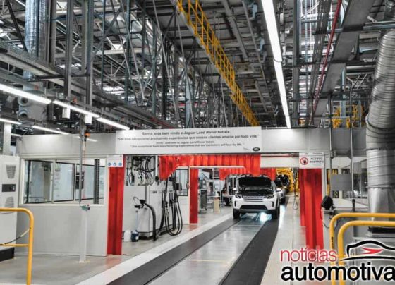 Interior de uma fábrica automotiva, com uma carroceria branca suspensa em uma linha de montagem com estruturas amarelas.