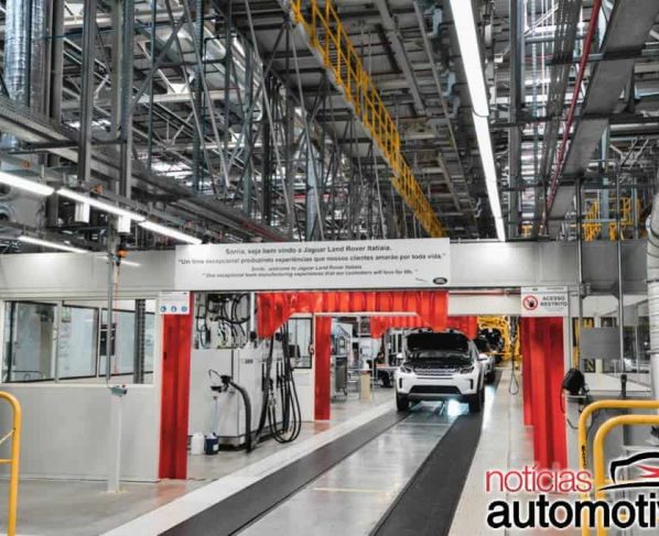 Interior de uma fábrica automotiva, com uma carroceria branca suspensa em uma linha de montagem com estruturas amarelas.